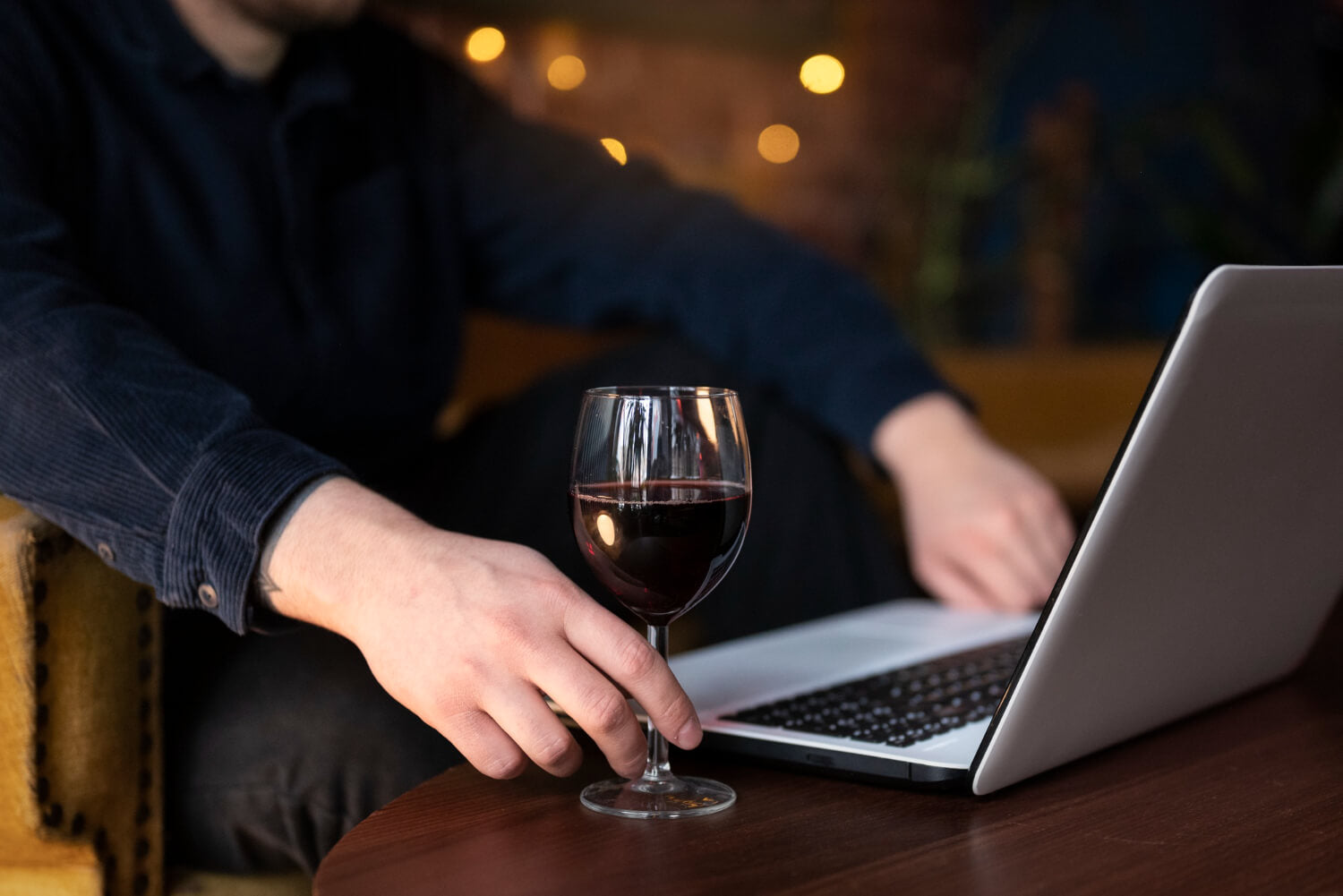 Person holding a glass of red wine next to a laptop on a wooden table.